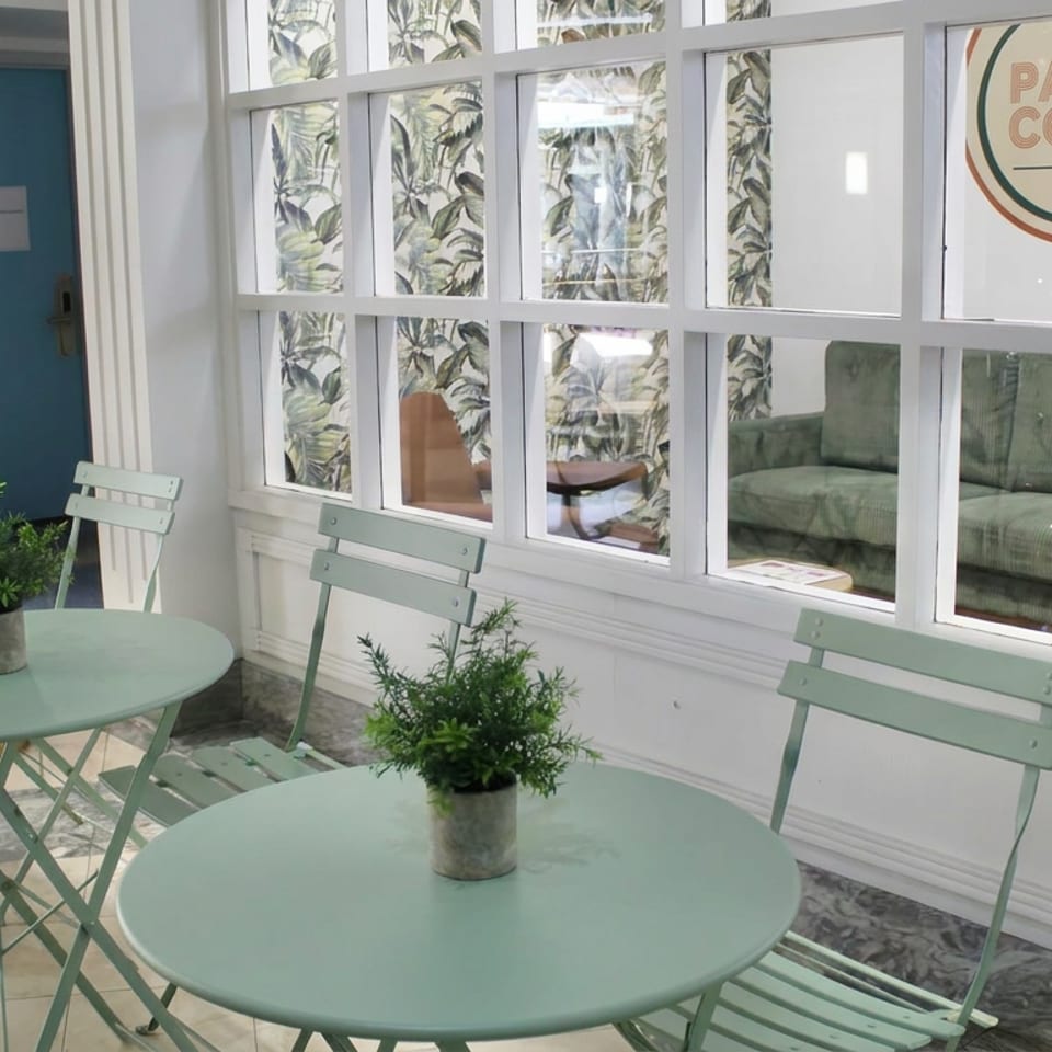 Bright Palm Court Cafe interior with mint-green tables, folding chairs, potted plants, and a lounge visible through glass windows.